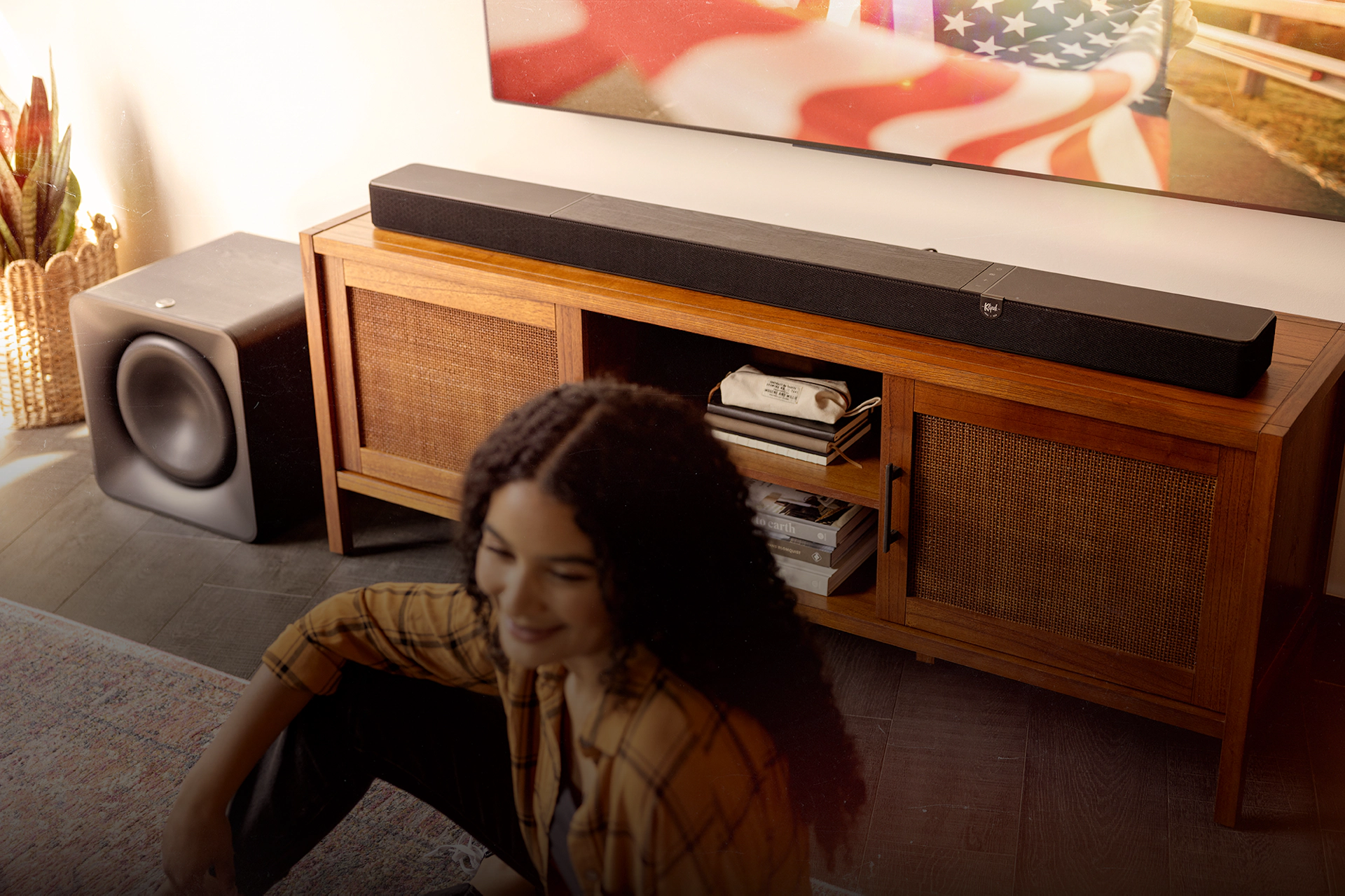 Klipsch Flexus CORE 300 BK and SUB 200 in focus in sunny living room with American flag on tv and woman in blurred foreground with overlay 2000x1333